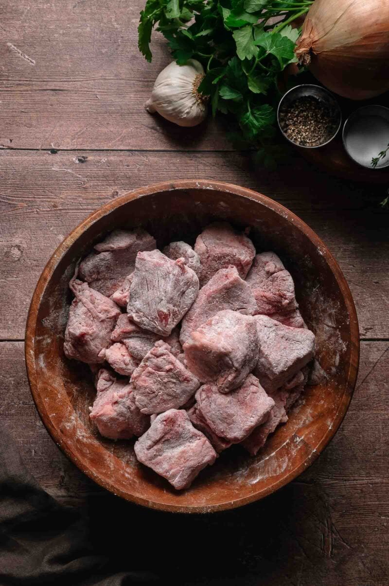 A wooden bowl filled with floured beef cubes sits on a wooden table, surrounded by garlic, an onion, fresh herbs, and small bowls of pepper.