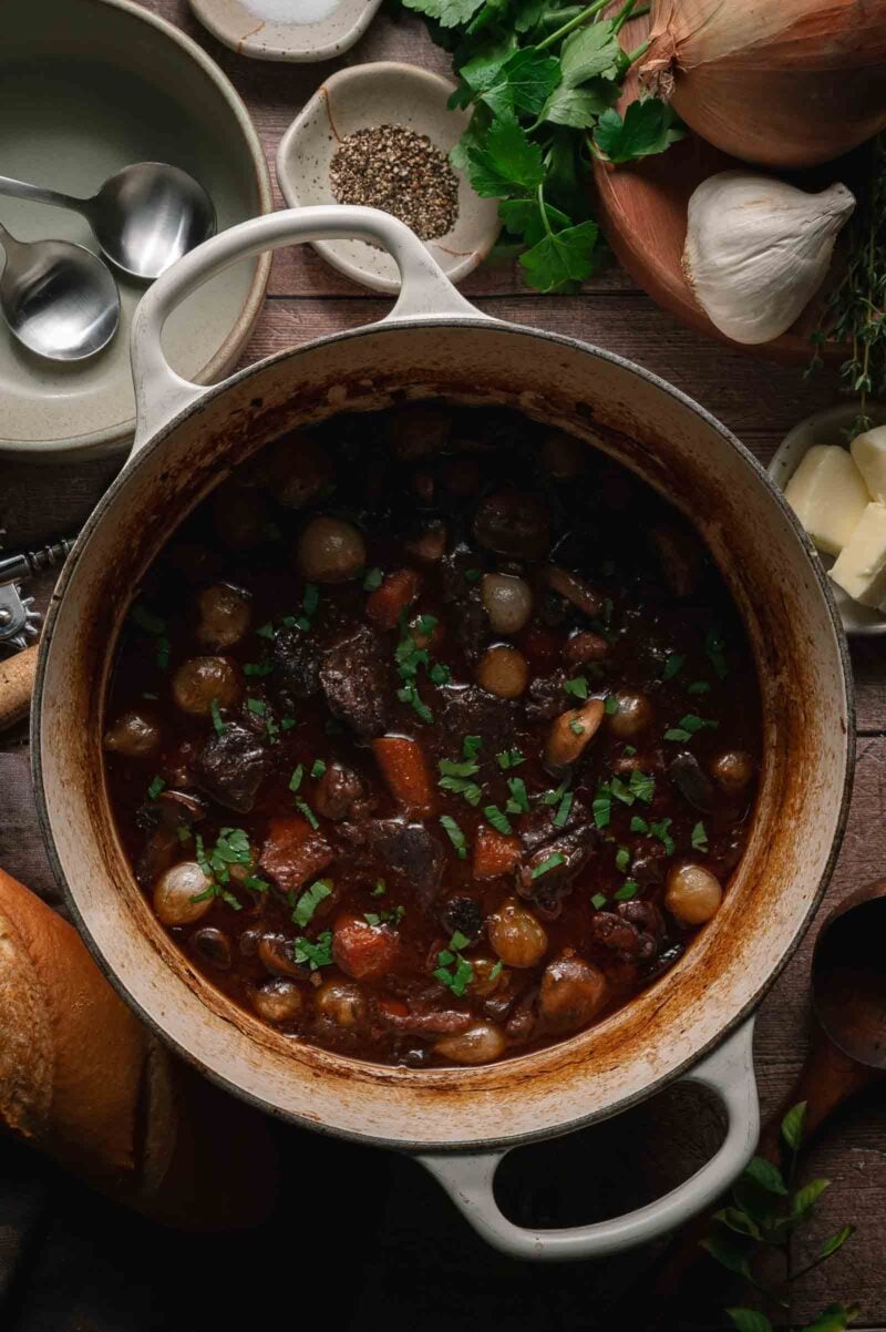 A pot of beef bourguignon with carrots, mushrooms, and pearl onions sits on a wooden table, surrounded by bread, garlic, onion, parsley, and seasoning.