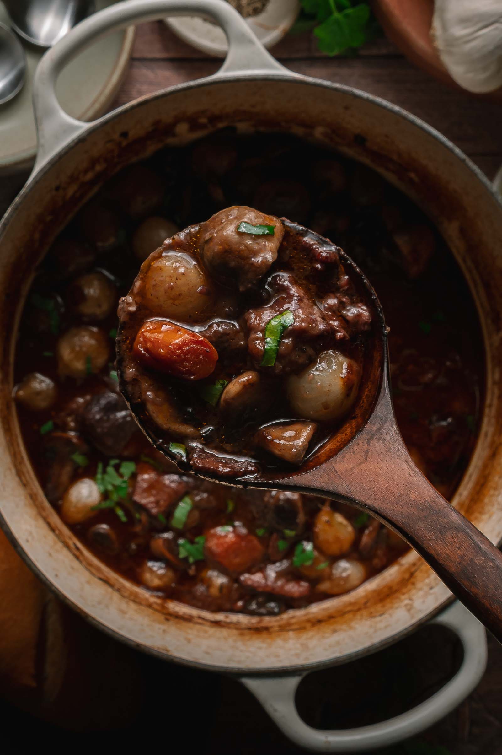 A ladle scoops beef bourguignon with pearl onions, carrots, and herbs from a round white Dutch oven.