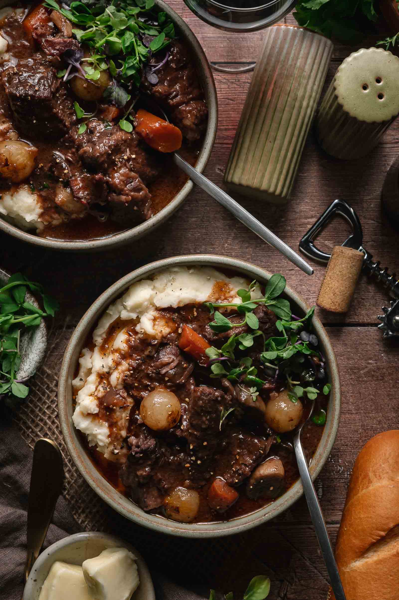 Two bowls of beef bourguignon with vegetables served over mashed potatoes, garnished with fresh herbs, on a wooden table with bread, butter, and a wine opener nearby.