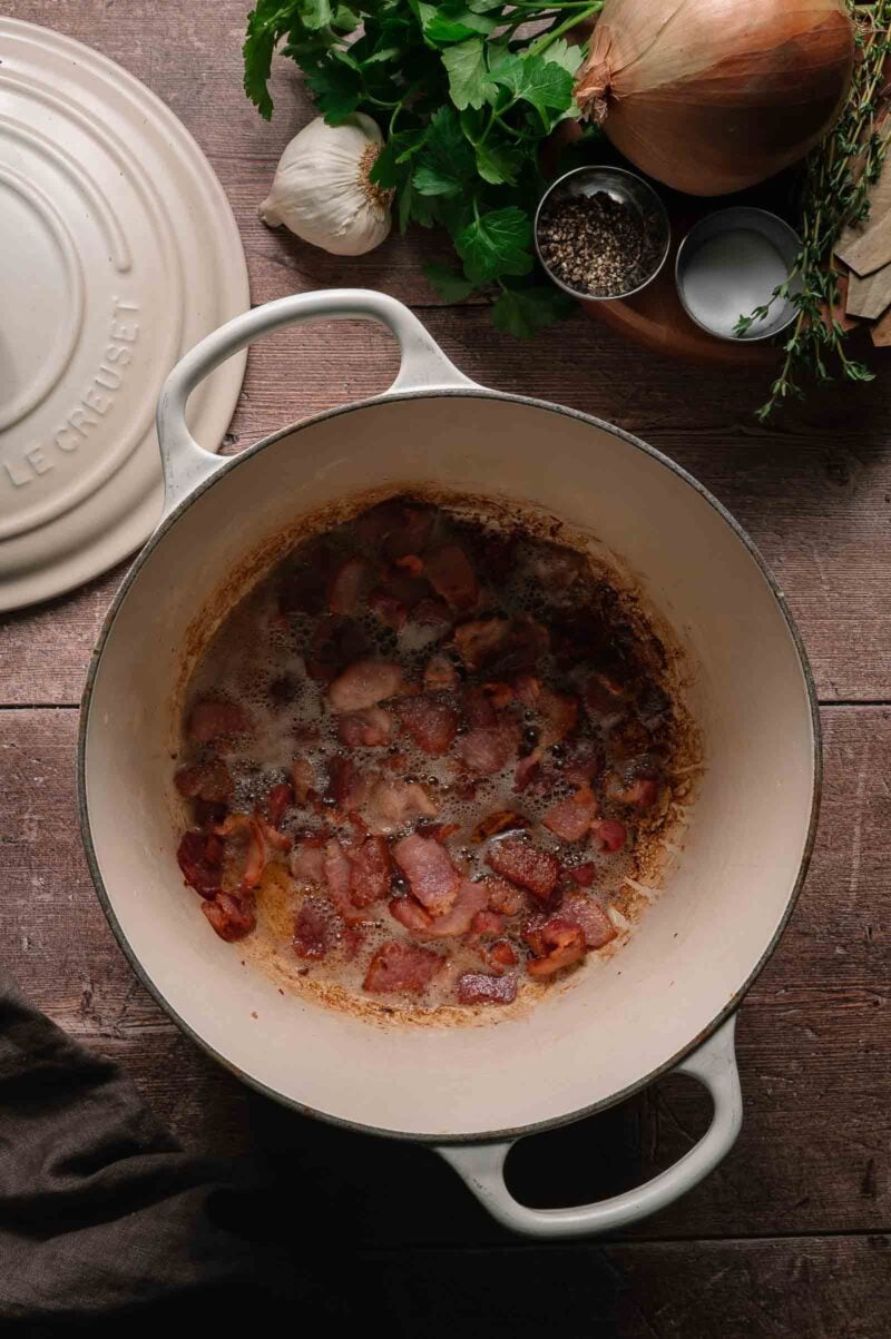 Chopped bacon is browning in a white Dutch oven; herbs, garlic, onion, salt, and pepper are arranged on the wooden surface next to it.