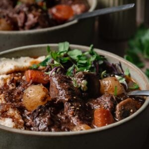 A bowl of beef bourguignon with carrots, potatoes, mushrooms, and fresh greens, served next to mashed potatoes.