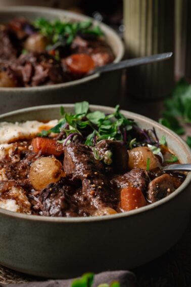 A bowl of beef bourguignon with carrots, potatoes, mushrooms, and fresh greens, served next to mashed potatoes.