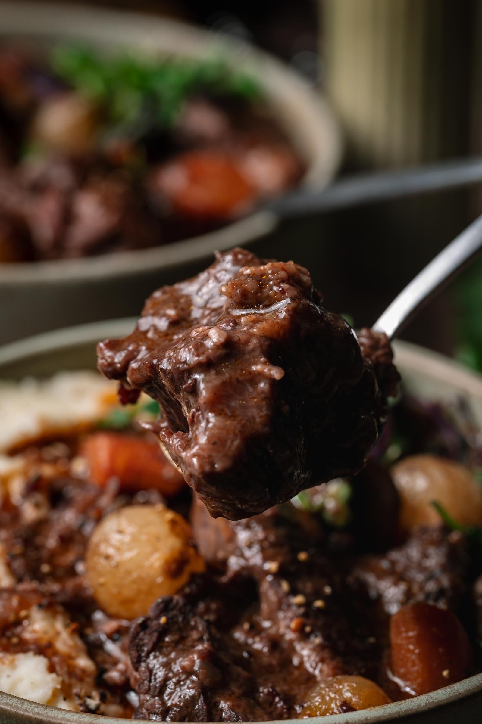 A close-up of a spoon holding a piece of beef bourguignon with pearl onions and carrots, with a bowl of the same stew in the background.