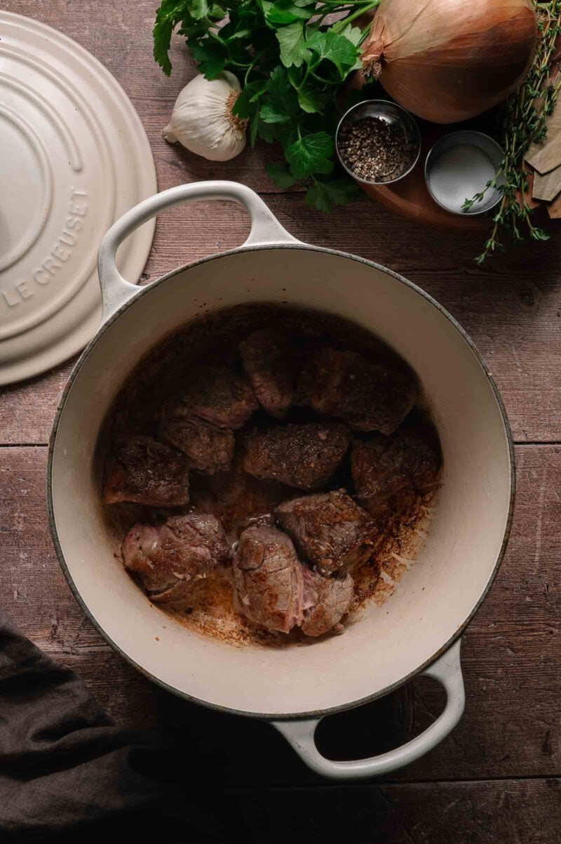 Chunks of browned beef chuck roast cooking in a white Dutch oven, surrounded by fresh herbs, an onion, garlic, salt, and pepper on a wooden surface.