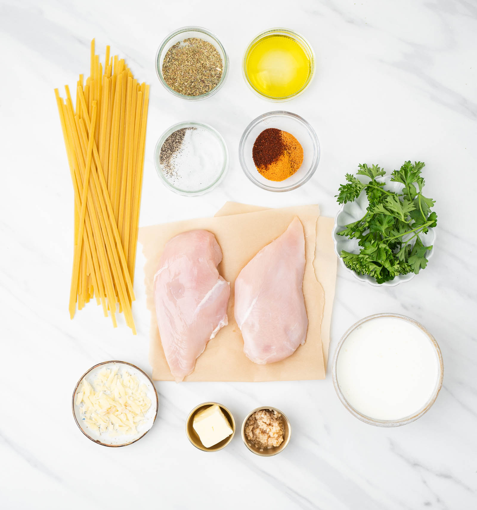 Uncooked pasta, raw chicken breasts, herbs, spices, oil, fresh parsley, shredded cheese, butter, minced garlic, and cream are arranged on a white surface.