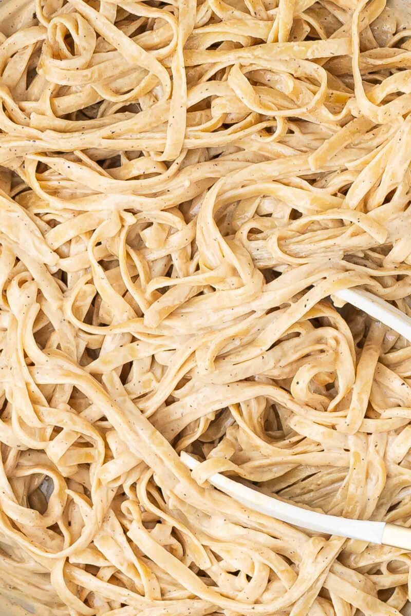 Close-up of fettuccine pasta coated in a creamy homemade alfredo sauce with black pepper, with a white serving utensil visible in the bottom right corner.