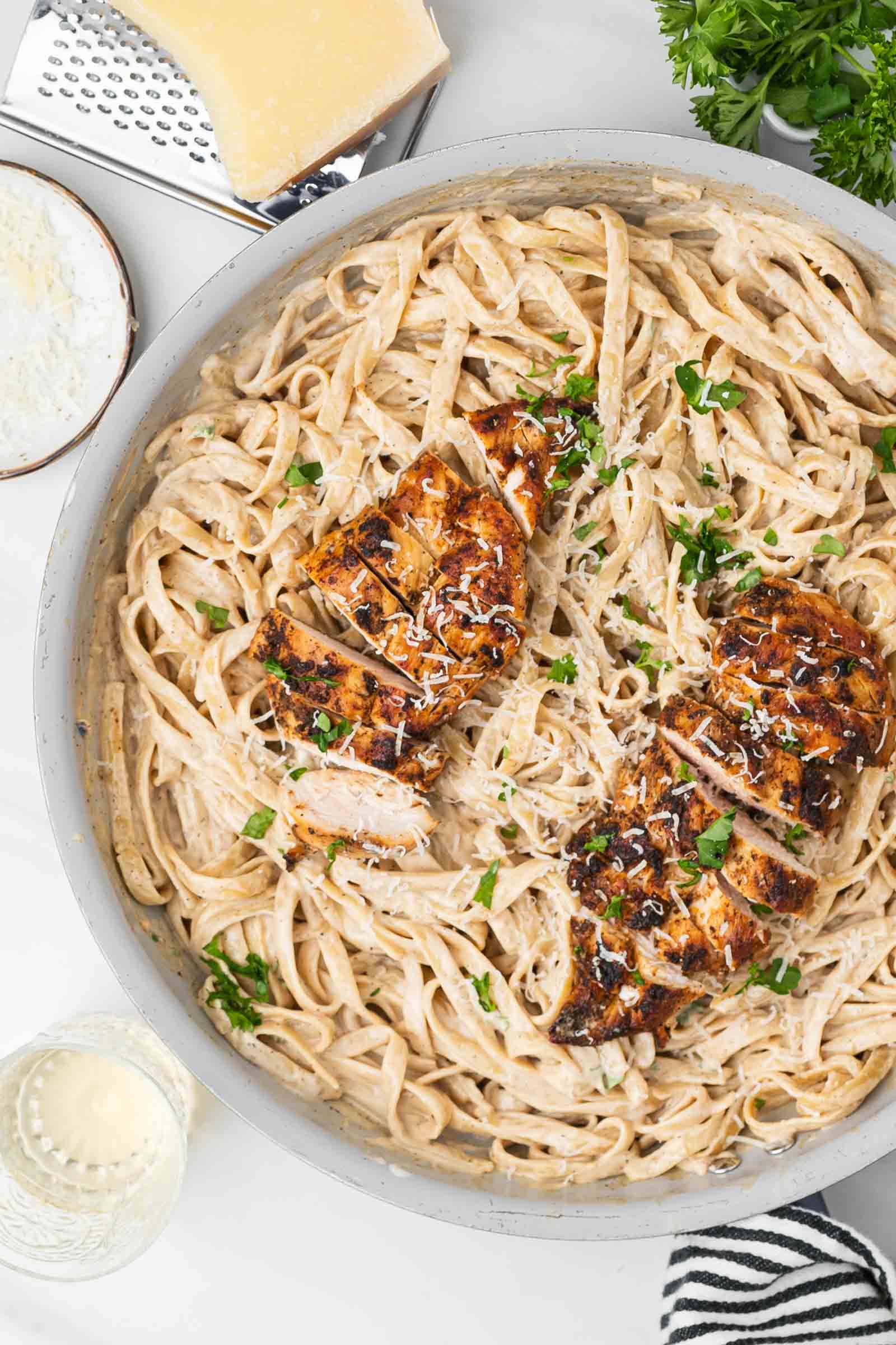 A pan of fettuccine alfredo topped with sliced blackened chicken breast, garnished with chopped parsley and grated cheese.