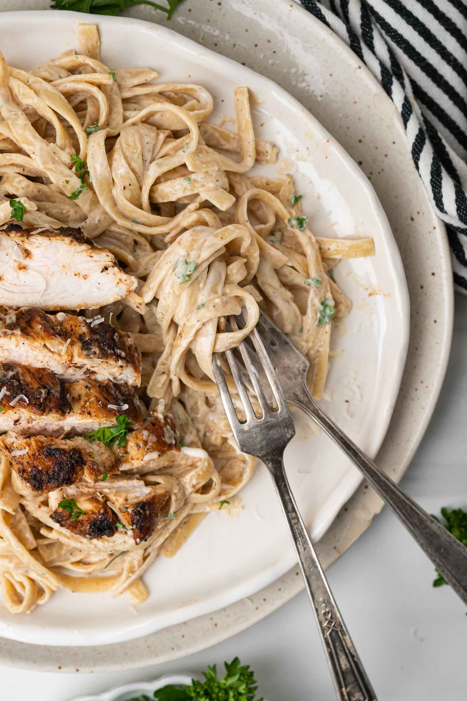 A plate of fettuccine Alfredo pasta with sliced blackened chicken, garnished with parsley, and two forks resting on the plate.