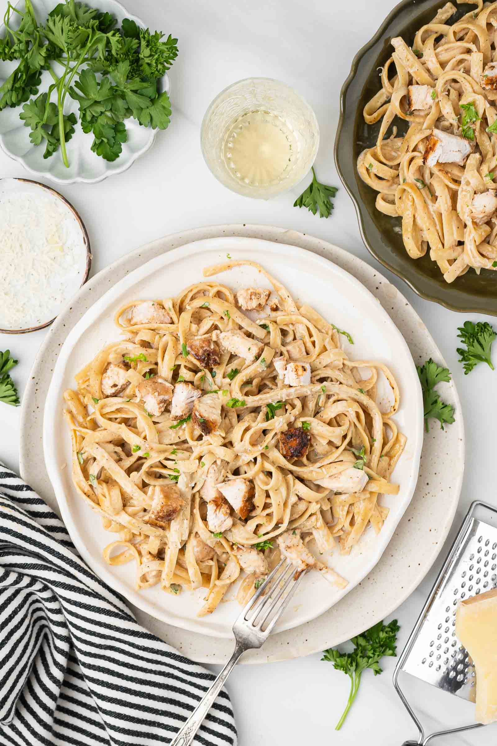 A plate of fettuccine pasta topped with blackened chicken pieces and garnished with parsley, next to a glass of white wine, grated cheese, and fresh herbs.