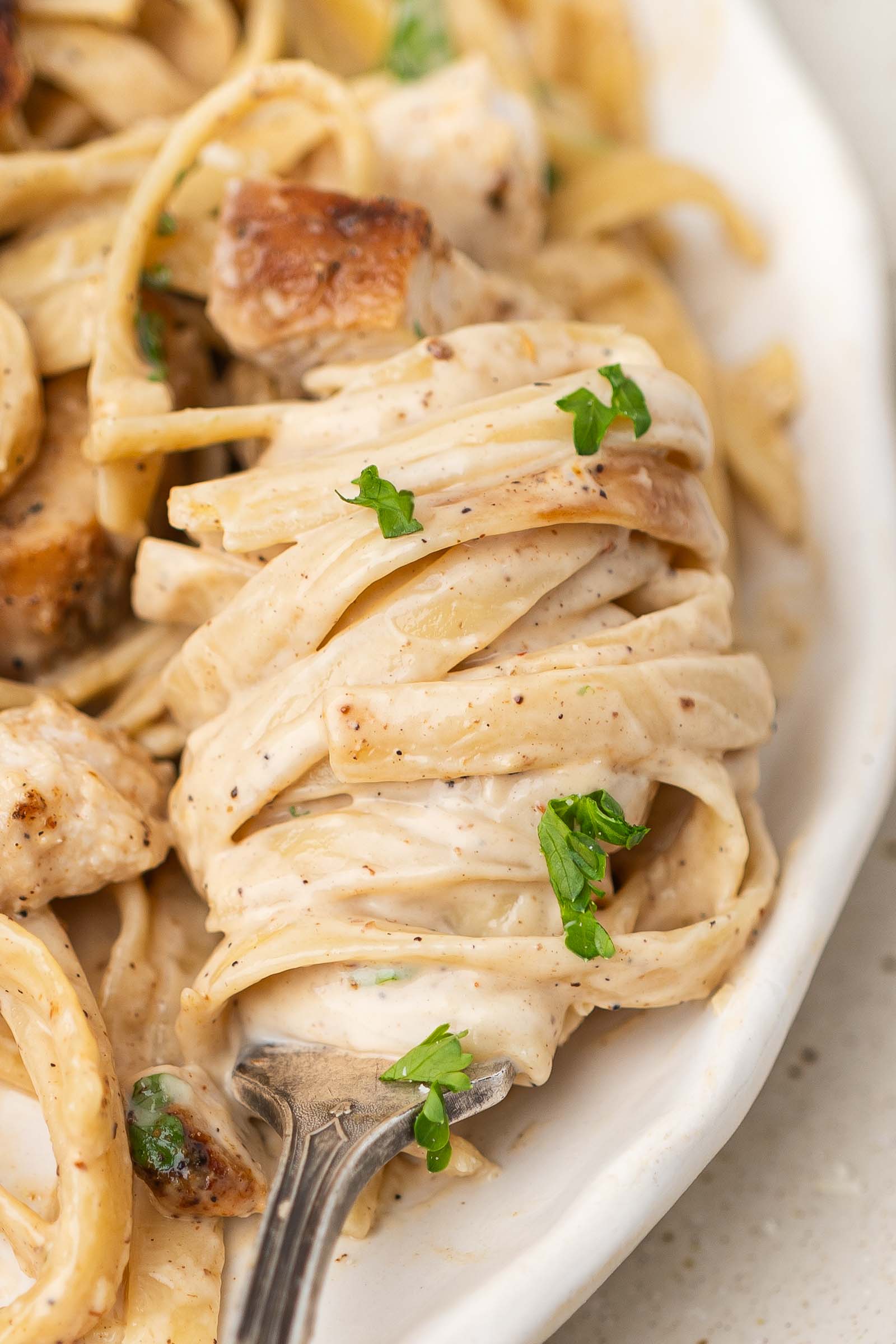 A fork twirls creamy fettuccine pasta with pieces of blackened chicken and parsley on a white plate.