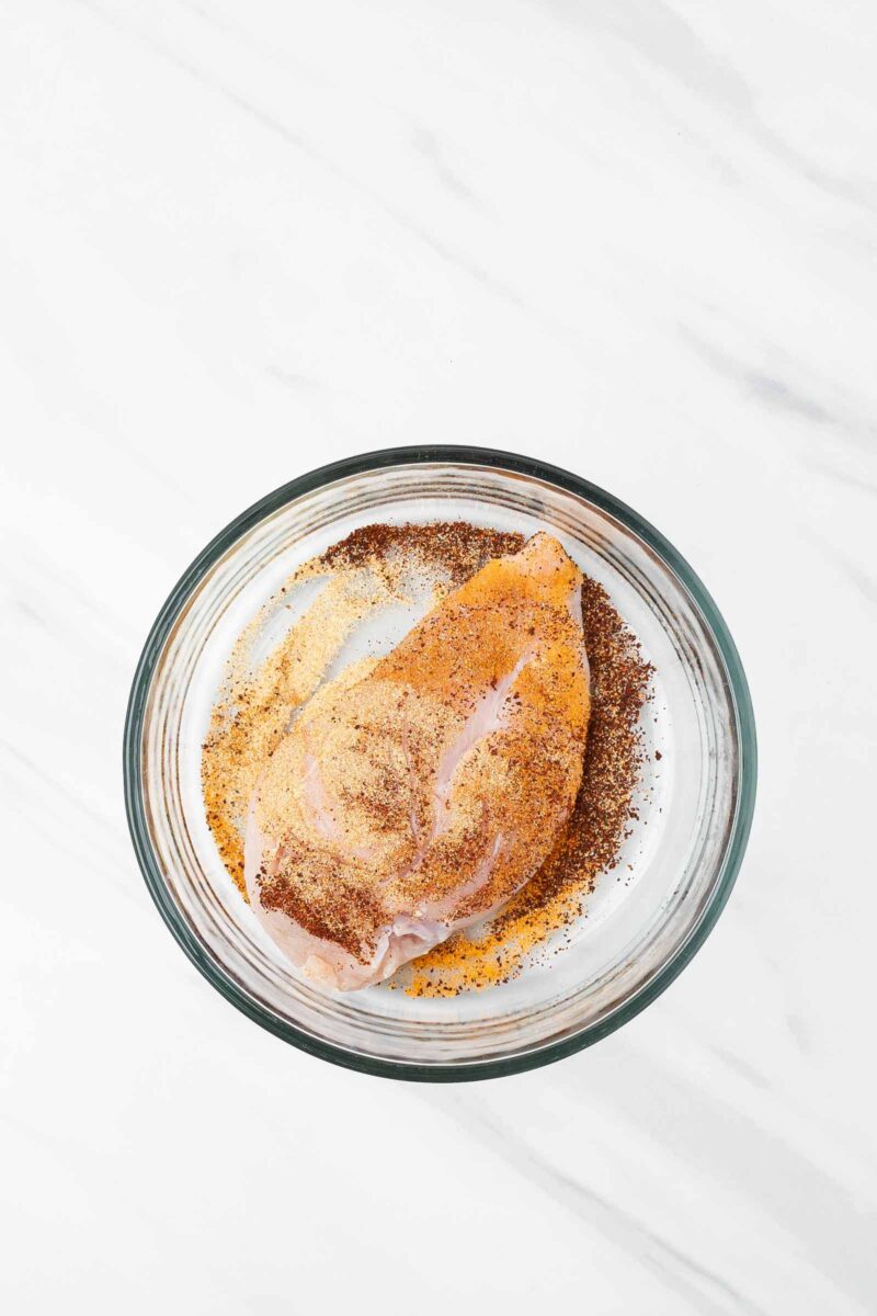 A raw chicken breast in a glass bowl is coated with assorted spices and seasoning on a white marble surface.