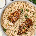 A skillet filled with fettuccine in a creamy sauce topped with sliced blackened chicken breast, garnished with grated cheese and chopped parsley.