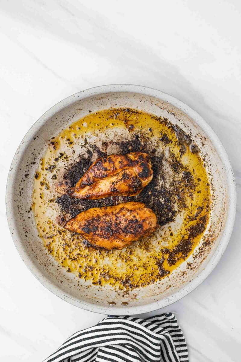 Two cooked, blackened chicken breasts in a white skillet with oil and seasoning residue, viewed from above on a marble surface. A striped towel is visible at the bottom edge.