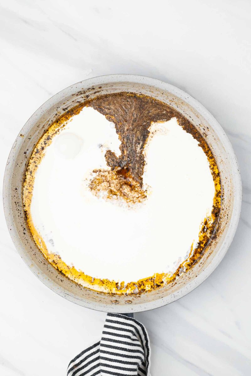 A frying pan on a white surface contains a mixture of cream or milk and brown seasonings, with a striped kitchen towel holding the handle.