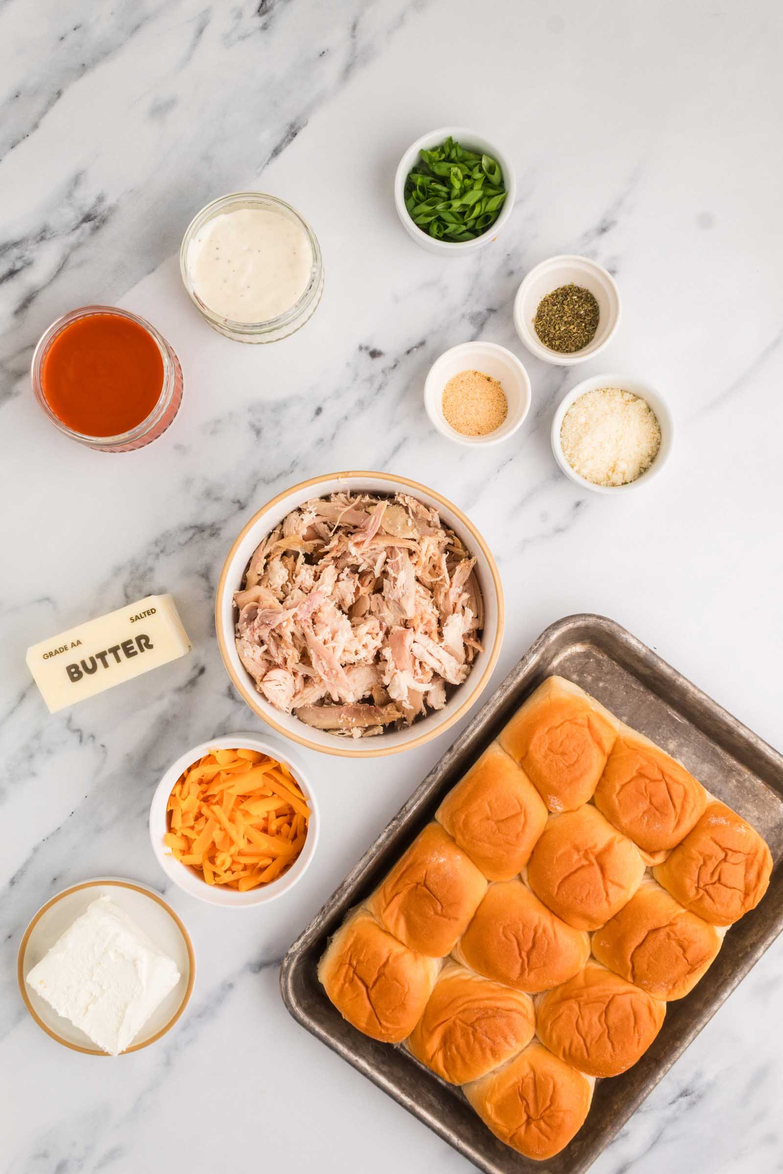 Various ingredients for making sliders are arranged on a marble surface, including shredded chicken, slider buns, cheese, butter, sauces, and seasonings.
