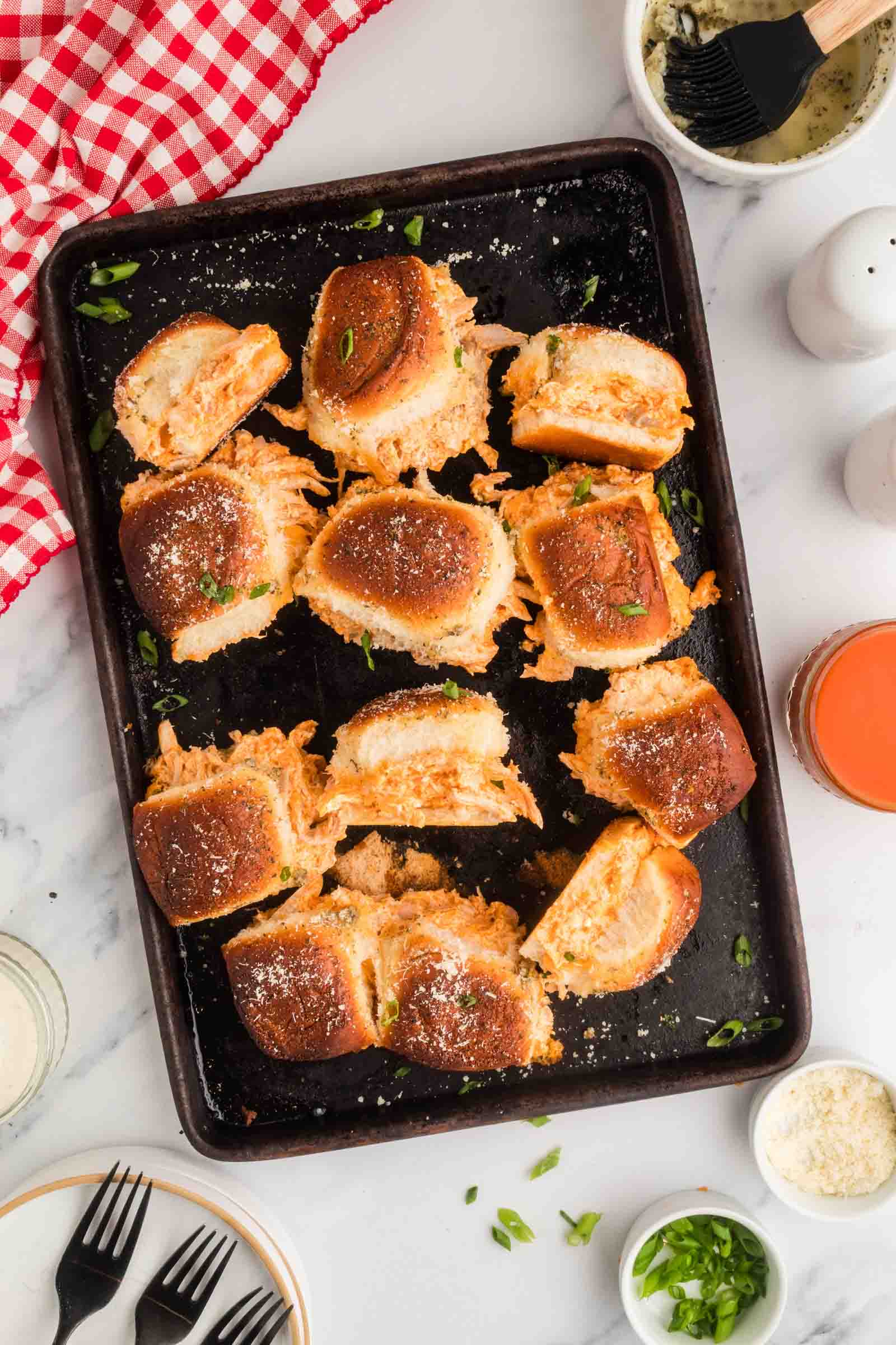 A baking tray with buffalo chicken baked sliders, some pulled apart, on a marble counter with a red checkered cloth and various small bowls and utensils nearby.