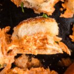 A close-up of a slider sandwich filled with shredded buffalo chicken on a dark surface, surrounded by similar sandwiches.
