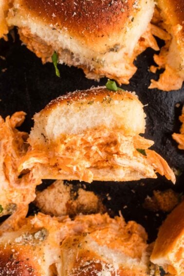 A close-up of a slider sandwich filled with shredded buffalo chicken on a dark surface, surrounded by similar sandwiches.