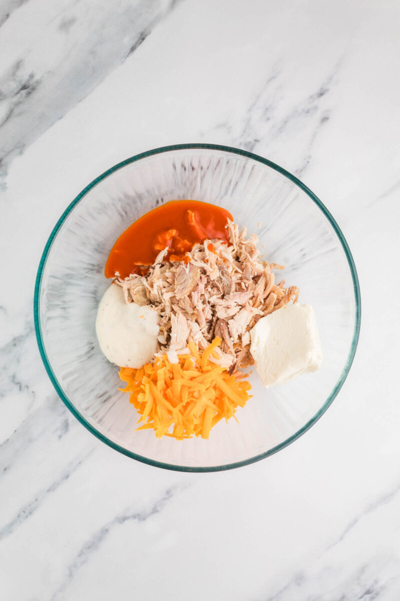 A glass bowl containing shredded chicken, shredded cheddar cheese, cream cheese, ranch dressing, and buffalo sauce on a marble surface.