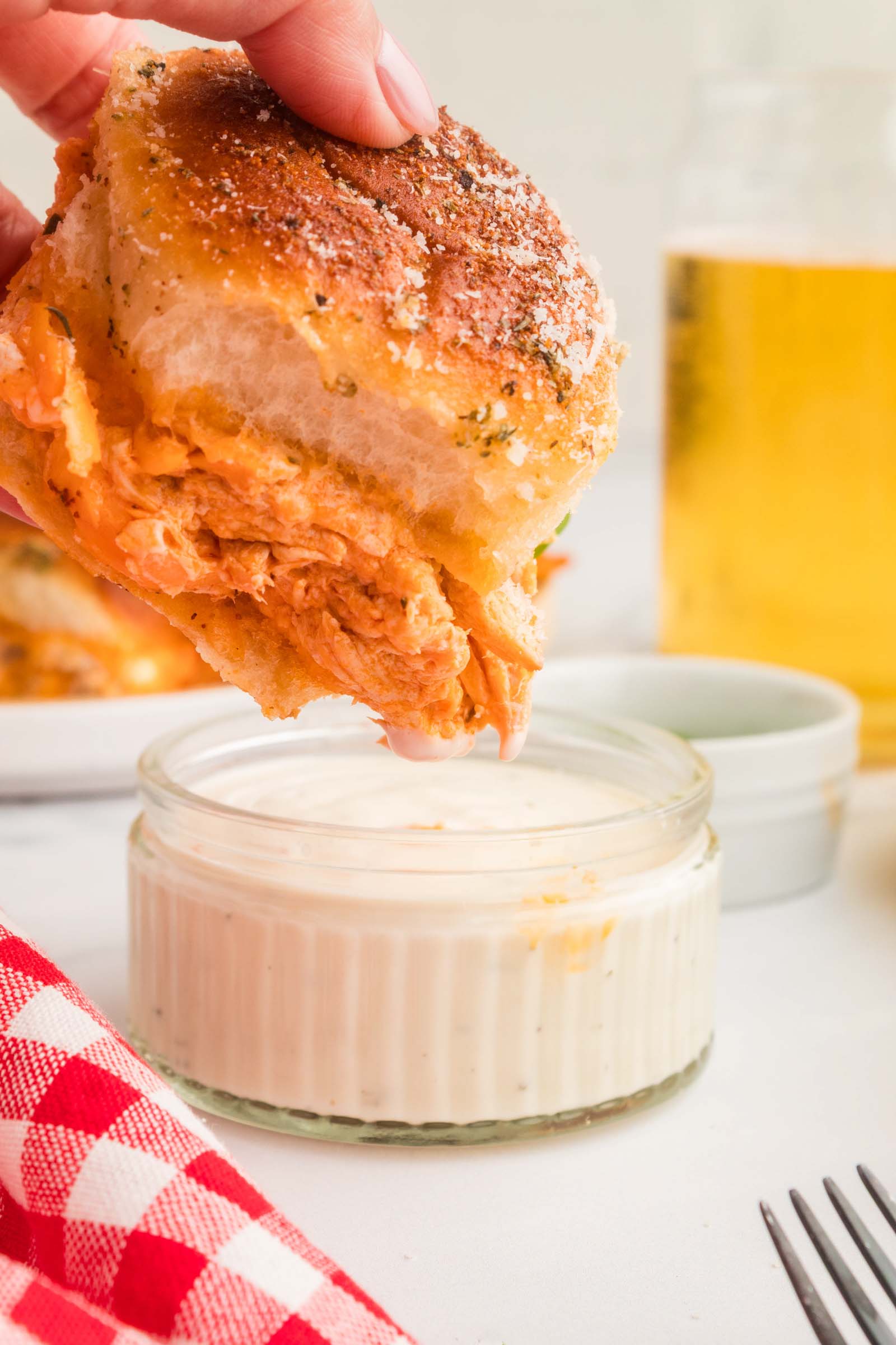 A hand dips a buffalo chicken slider into a small glass jar of ranch dressing, with a drink, napkin, and fork nearby.