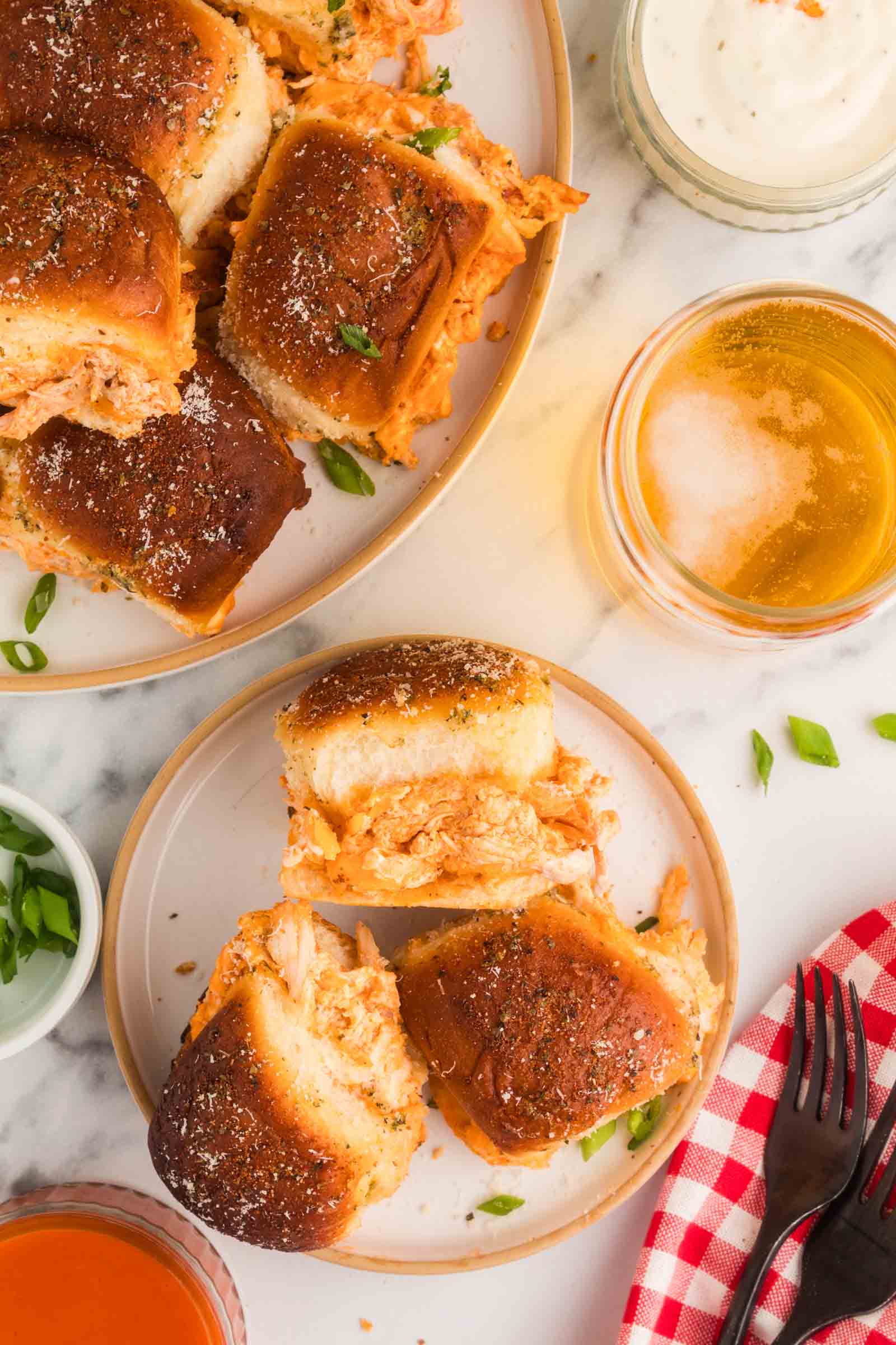 Two plates with buffalo chicken sliders on hawaiian slider rolls, garnished with herbs, surrounded by small bowls of dipping sauces, sliced green onions, and drinks on a marble surface.