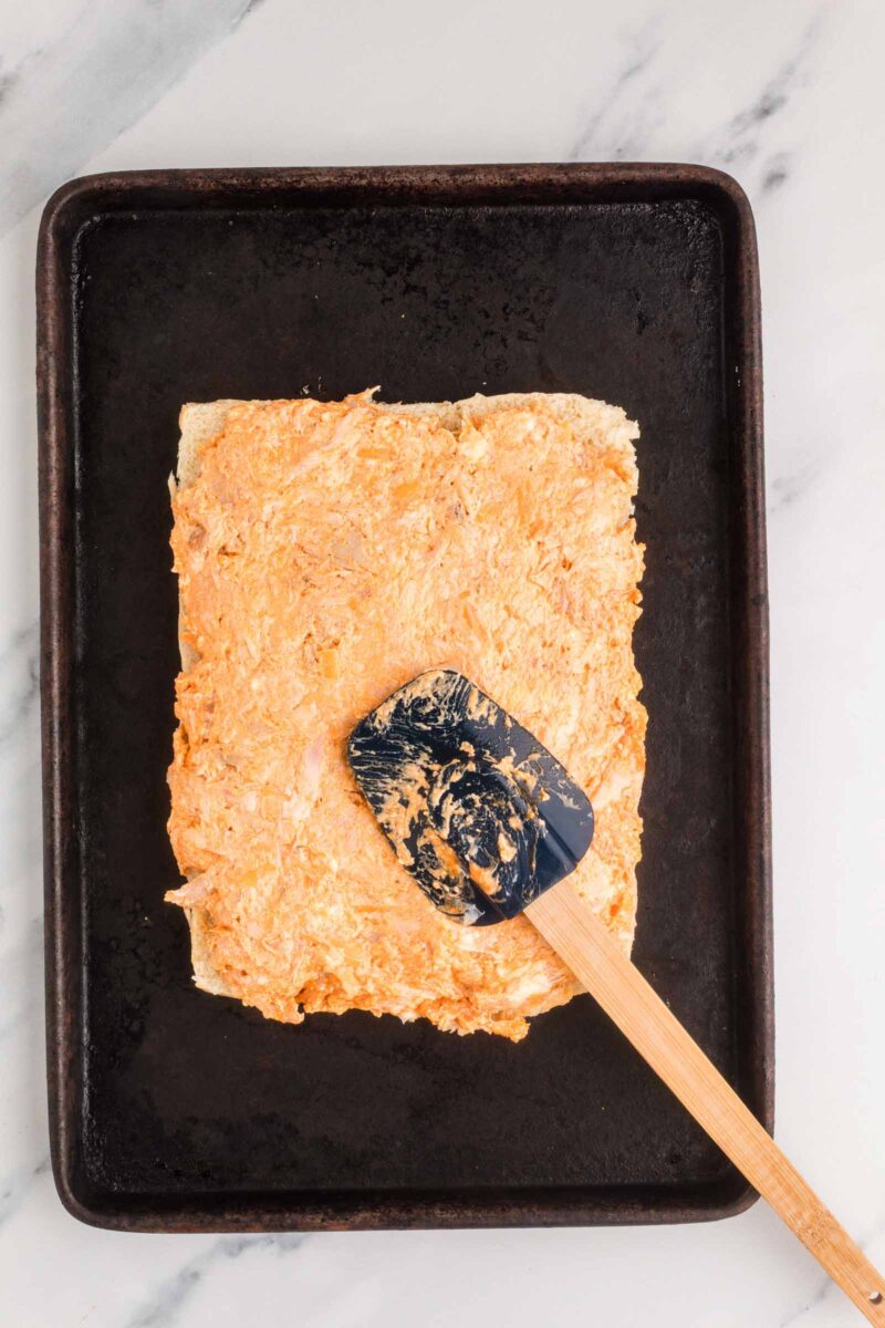 A rectangular piece of bread spread with a creamy buffalo chicken mixture sits on a black baking sheet, with a spatula resting on top.