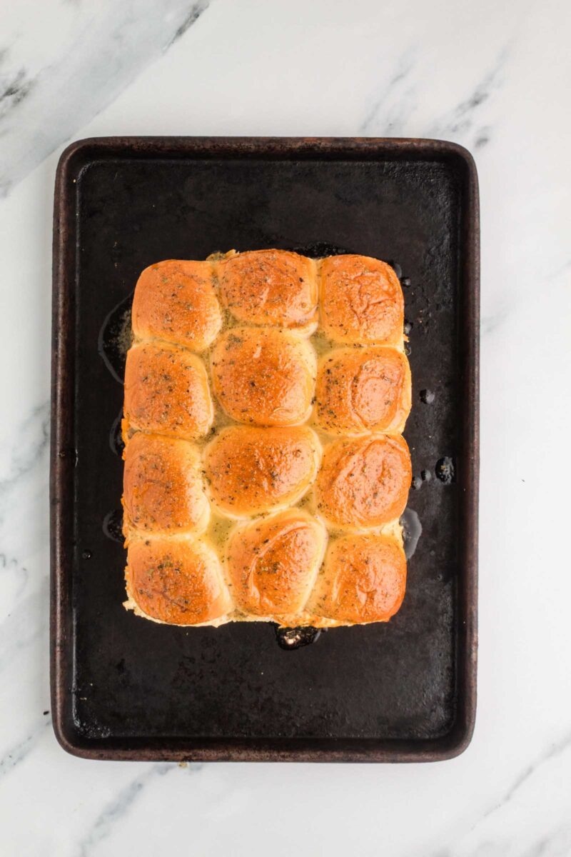 A batch of twelve slider buns sits on a baking tray, buttered and sprinkled with seasoning ready to go into the oven, on a white marble surface.