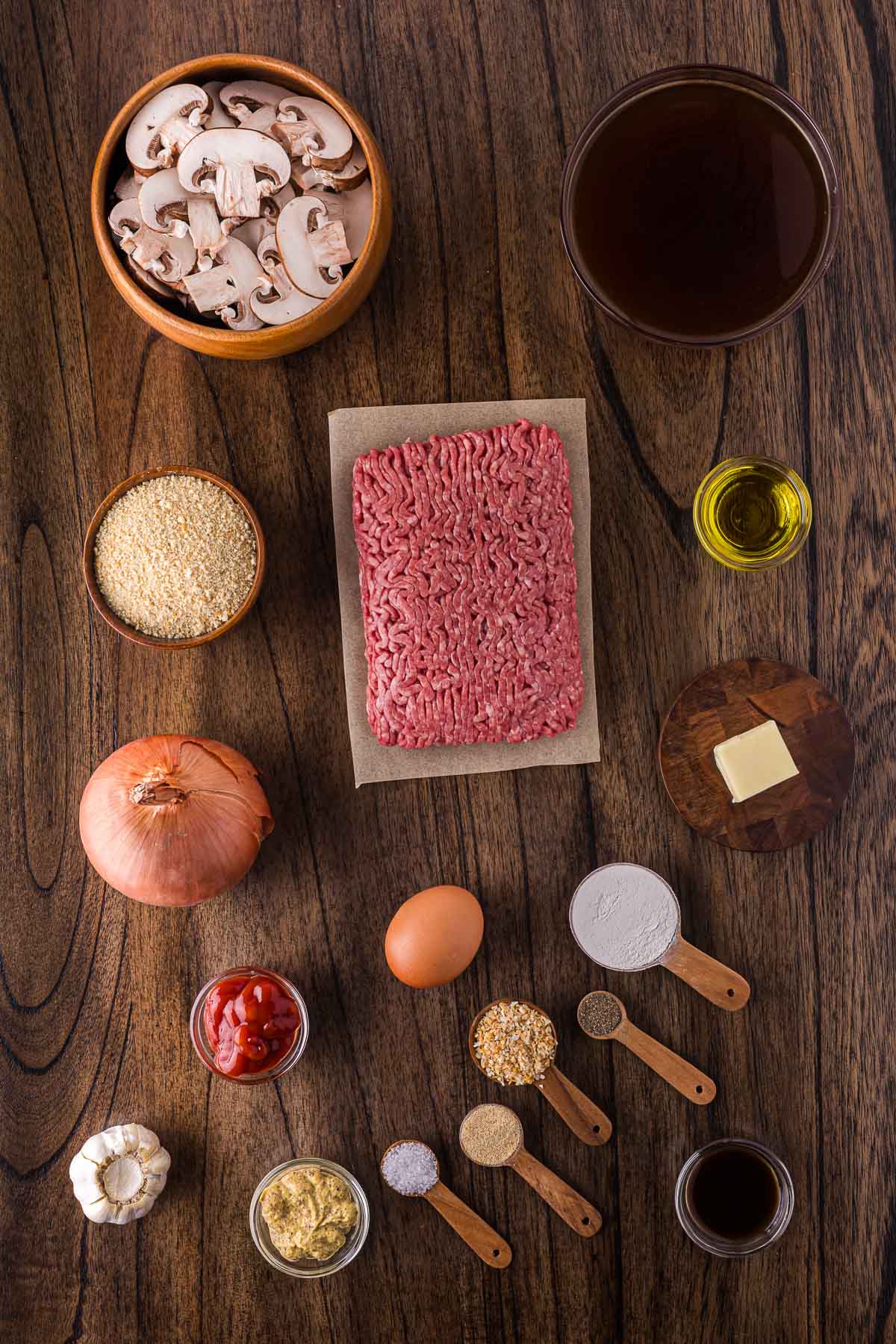 Ingredients for a Salisbury steaks recipe arranged on a wooden surface, including ground beef, sliced mushrooms, onion, egg, spices, broth, oil, butter, tomato paste, and breadcrumbs.
