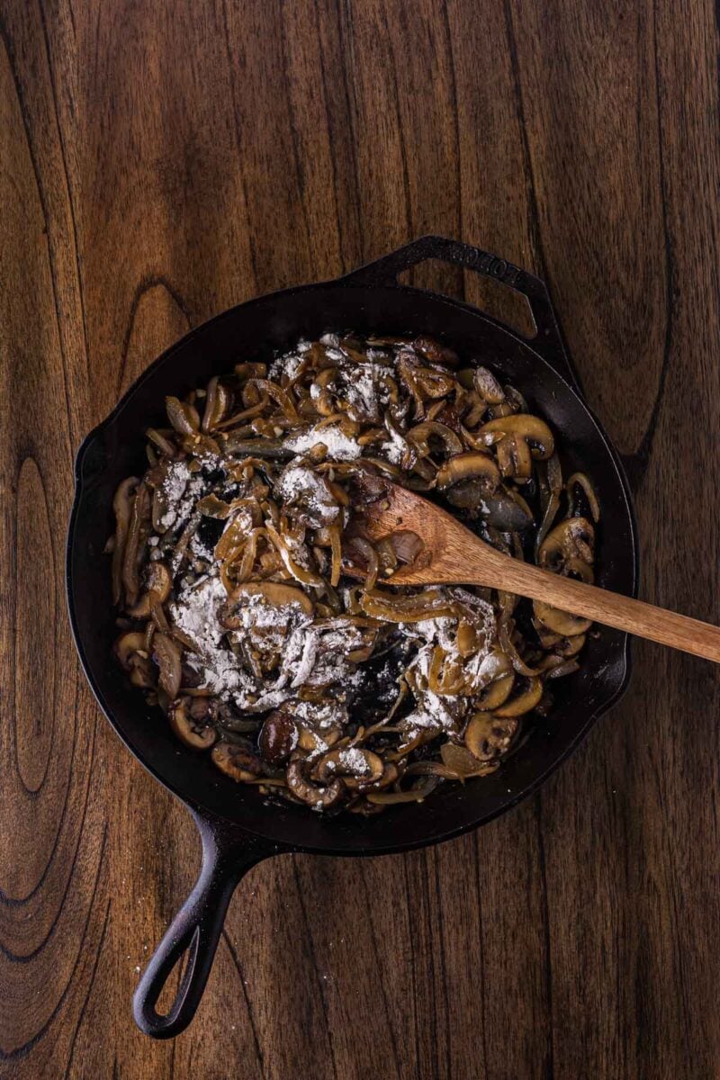 A cast iron skillet with sliced mushrooms and onions being stirred with a wooden spoon, topped with a layer of flour, on a wooden surface.