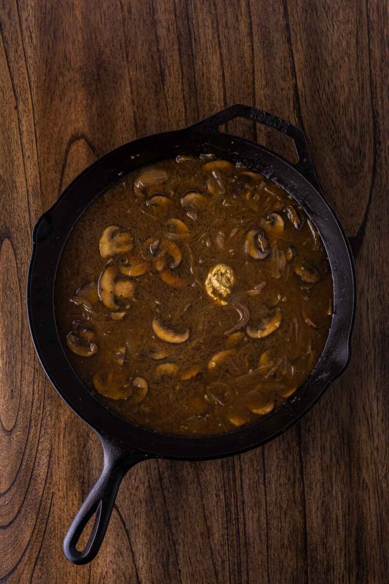 A cast iron skillet filled with mushroom and onion gravy sits on a wooden surface.