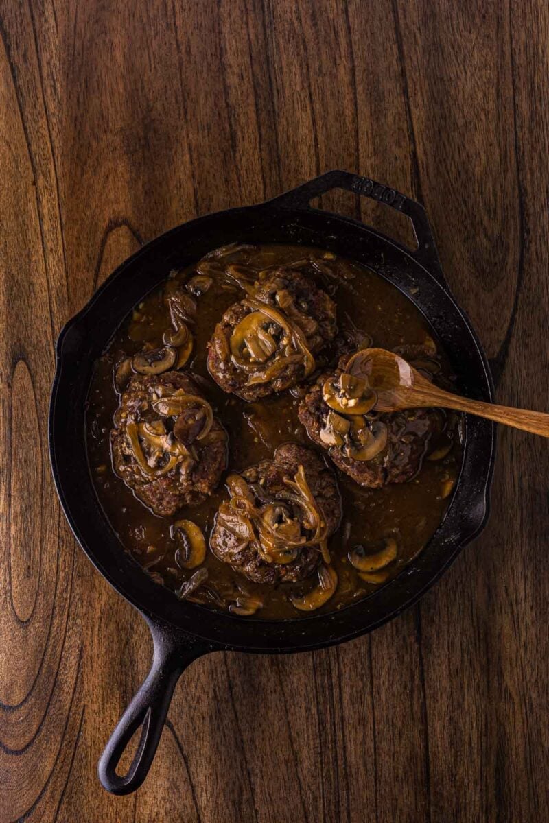 Cast iron skillet with hamburger steaks topped with sautéed onions and mushrooms in brown gravy, on a wooden surface. A wooden spoon rests on the skillet.