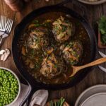 A cast iron skillet with four cooked Salisbury steaks in brown gravy with onions, surrounded by mashed potatoes, green peas, mushrooms, herbs, and tableware on a wooden surface.