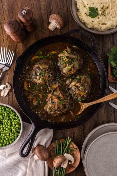 A cast iron skillet with four cooked Salisbury steaks in brown gravy with onions, surrounded by mashed potatoes, green peas, mushrooms, herbs, and tableware on a wooden surface.