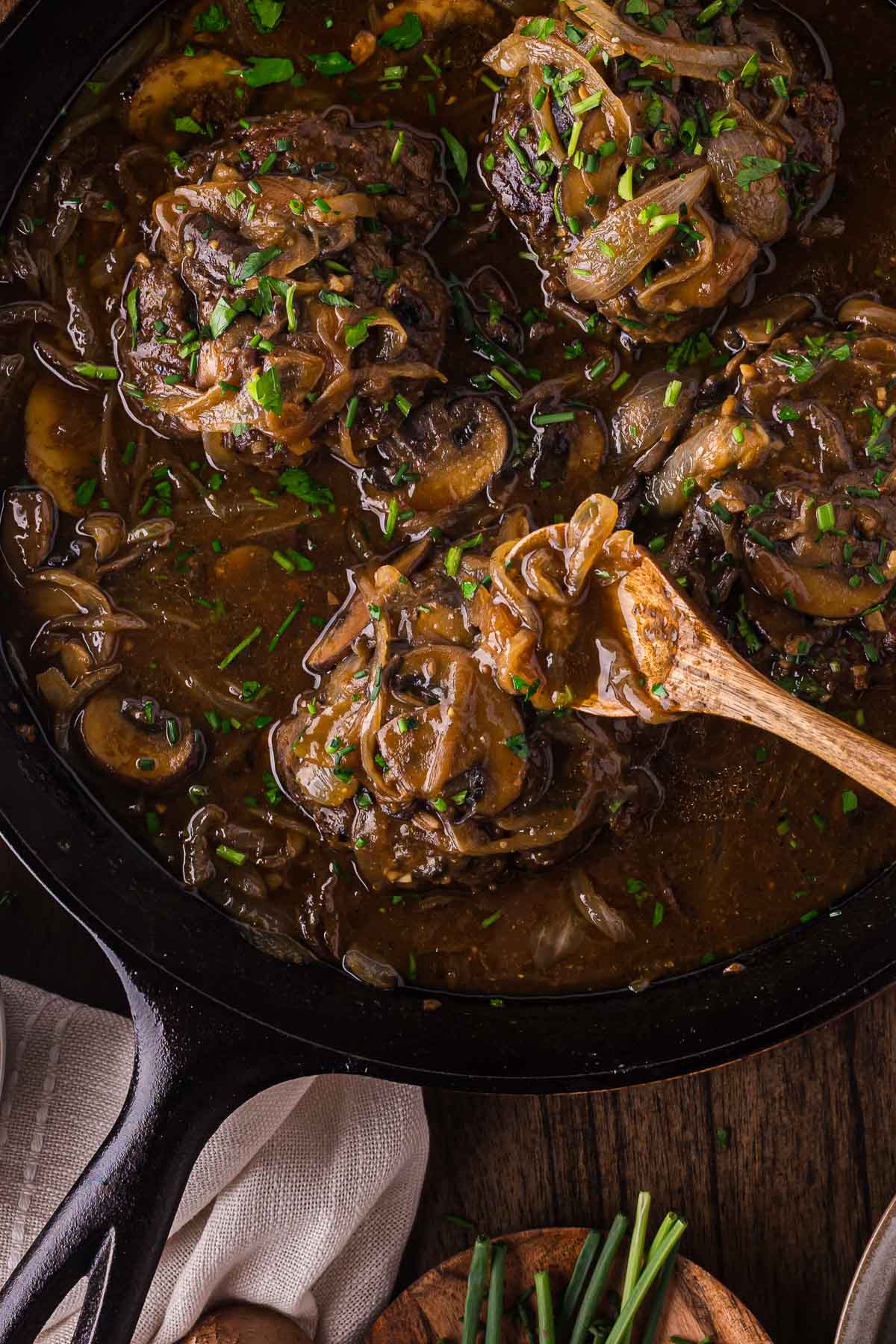A cast iron skillet filled with hamburger steaks topped with sautéed onions and mushrooms in brown gravy, garnished with chopped herbs.