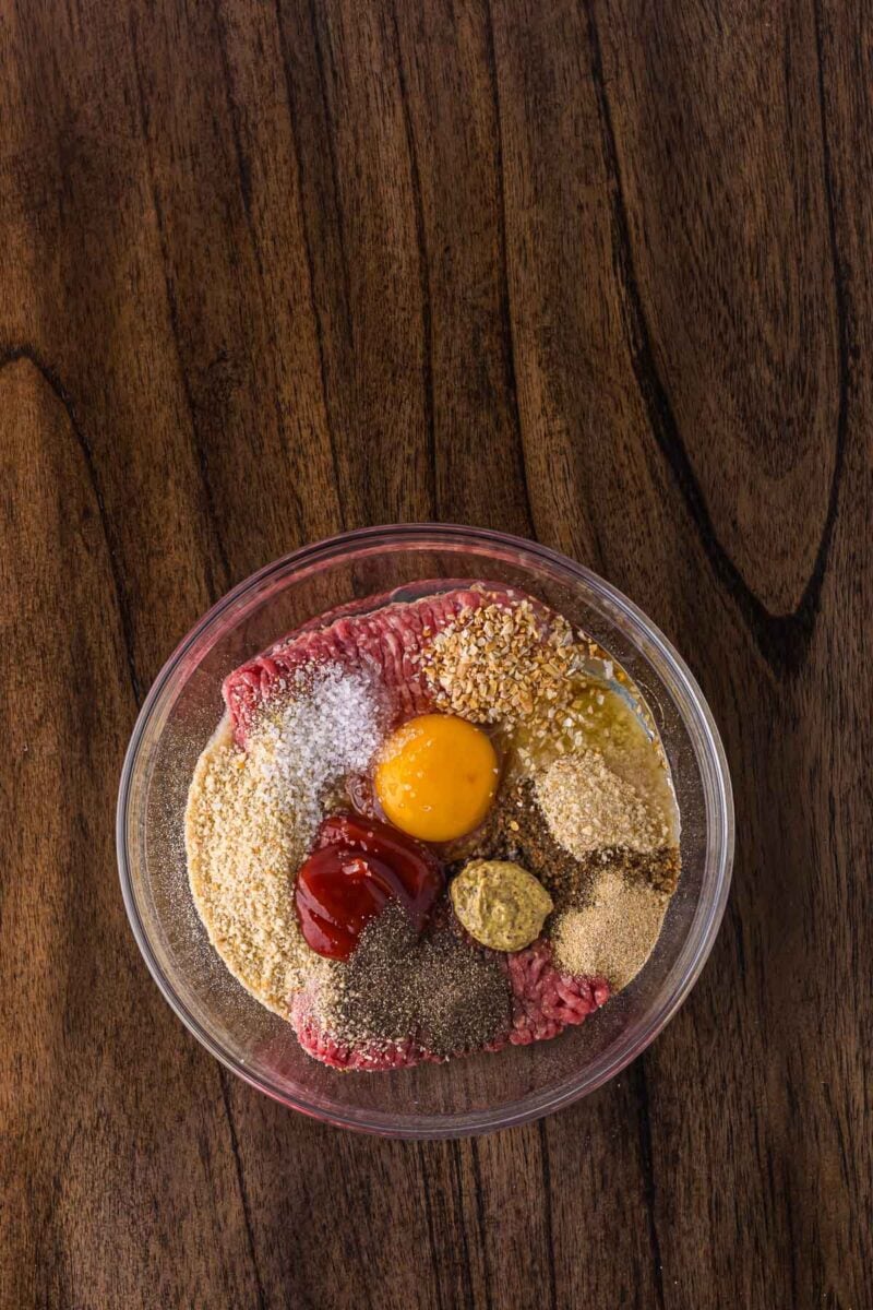 A glass bowl containing ground beef, an egg yolk, ketchup, mustard, spices, breadcrumbs, and seasonings on a wooden surface.