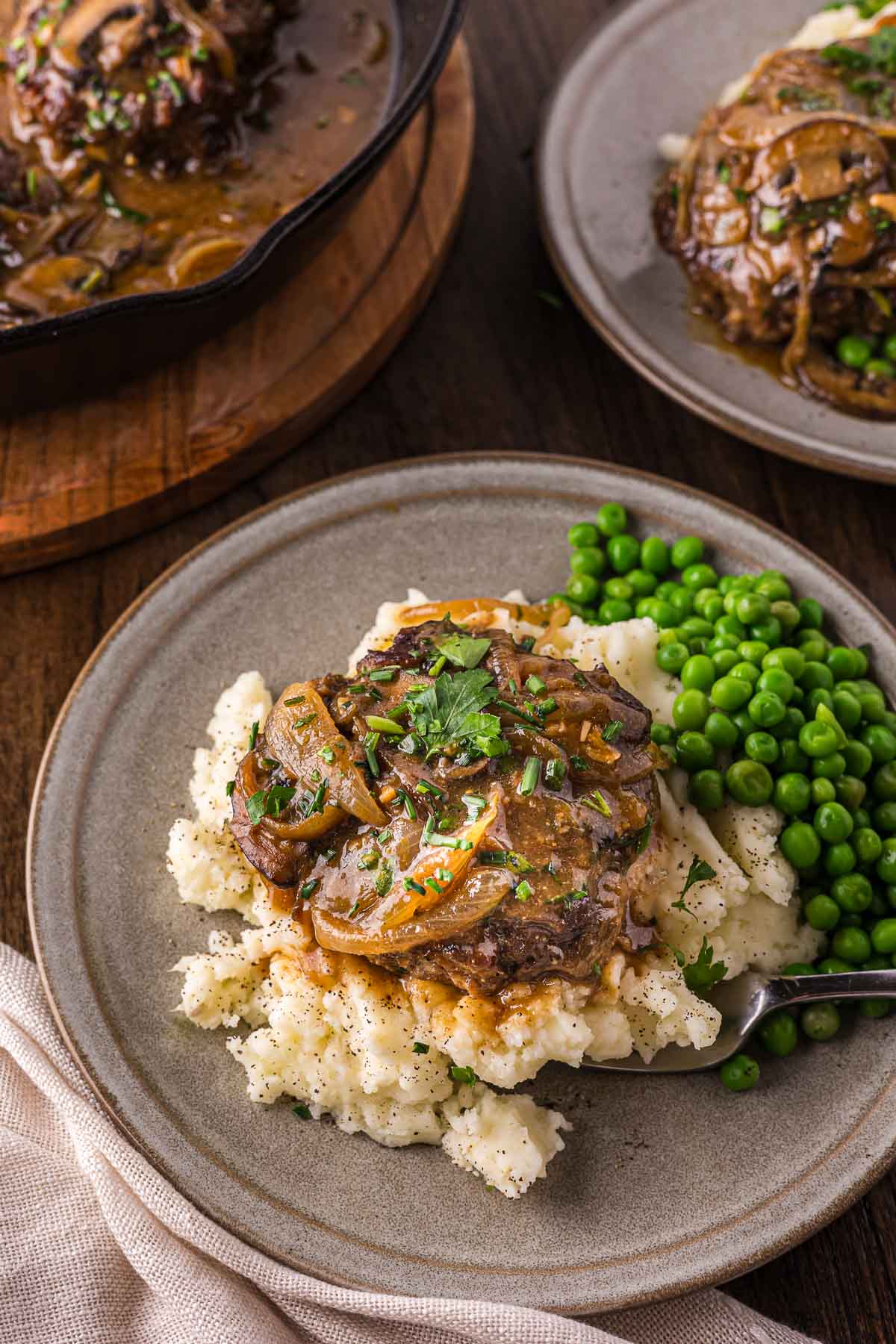 A plate of mashed potatoes topped with a Salisbury steak in onion and mushroom gravy, served with green peas on the side.