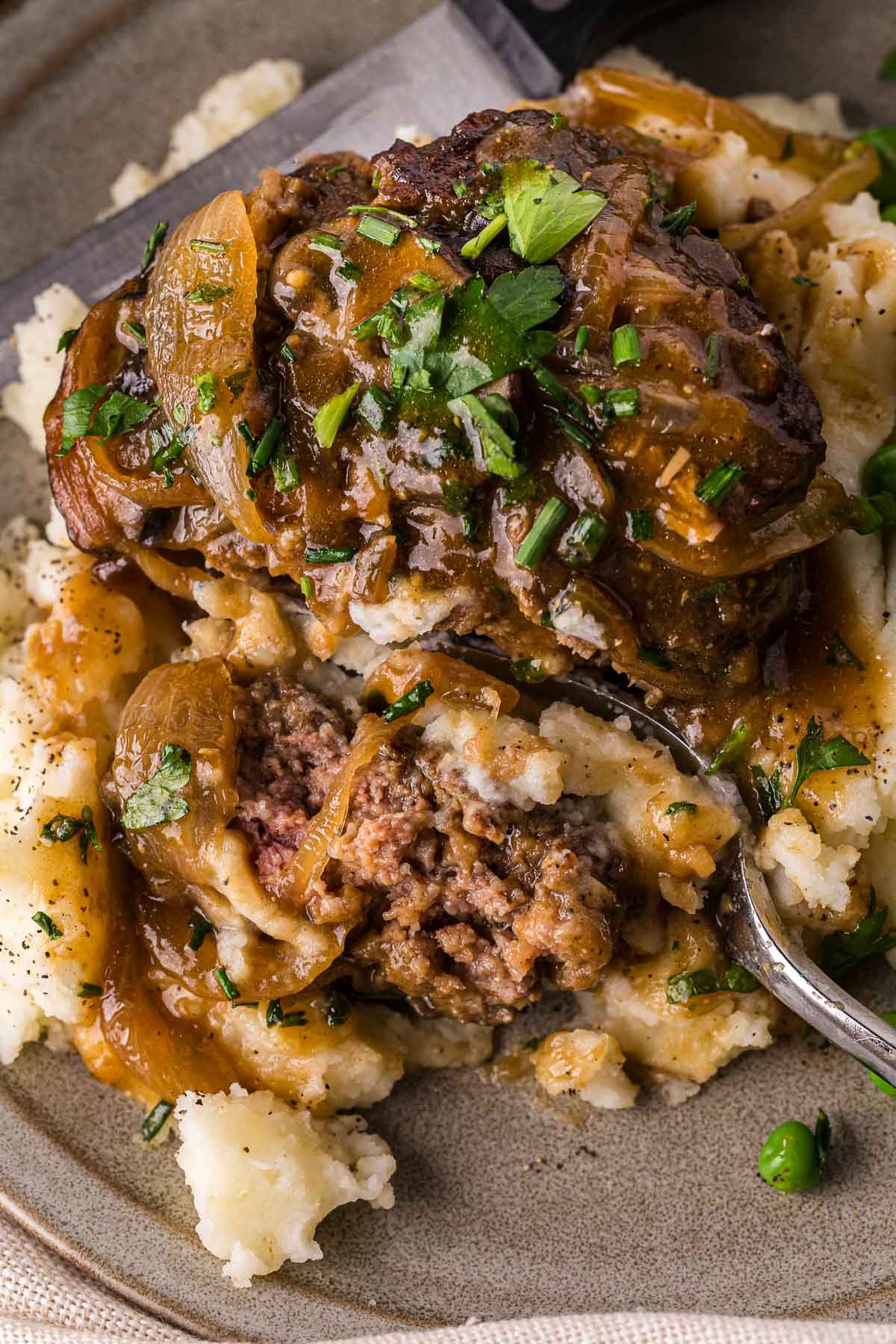 A serving of Salisbury steak with brown onion gravy sits on mashed potatoes, garnished with chopped parsley, on a plate. A fork holds a bite-sized piece of the dish.
