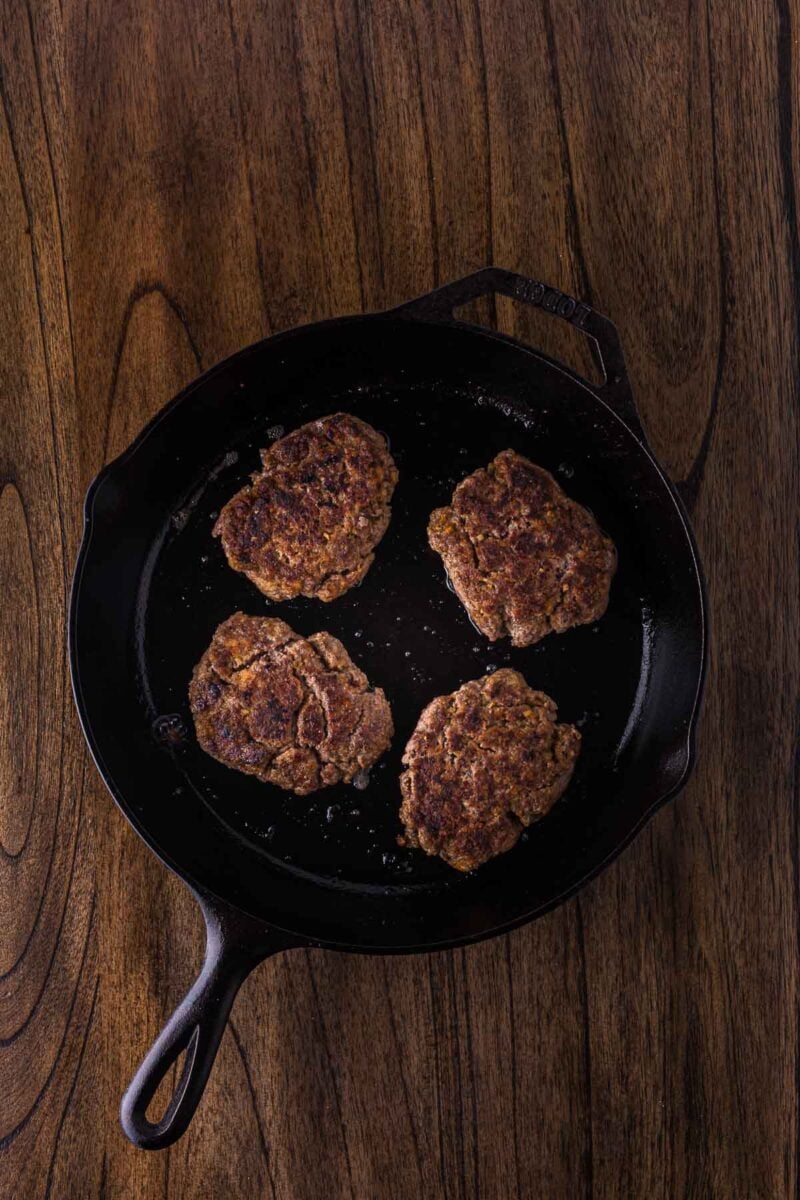 Four cooked burger steaks in a cast iron skillet on a wooden surface, viewed from above.