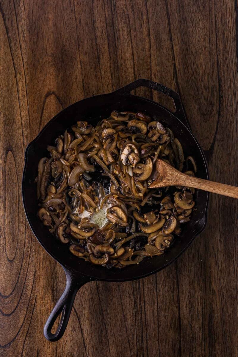 A cast iron skillet filled with sautéed mushrooms and onions sits on a wooden surface, with a wooden spoon resting inside the pan.