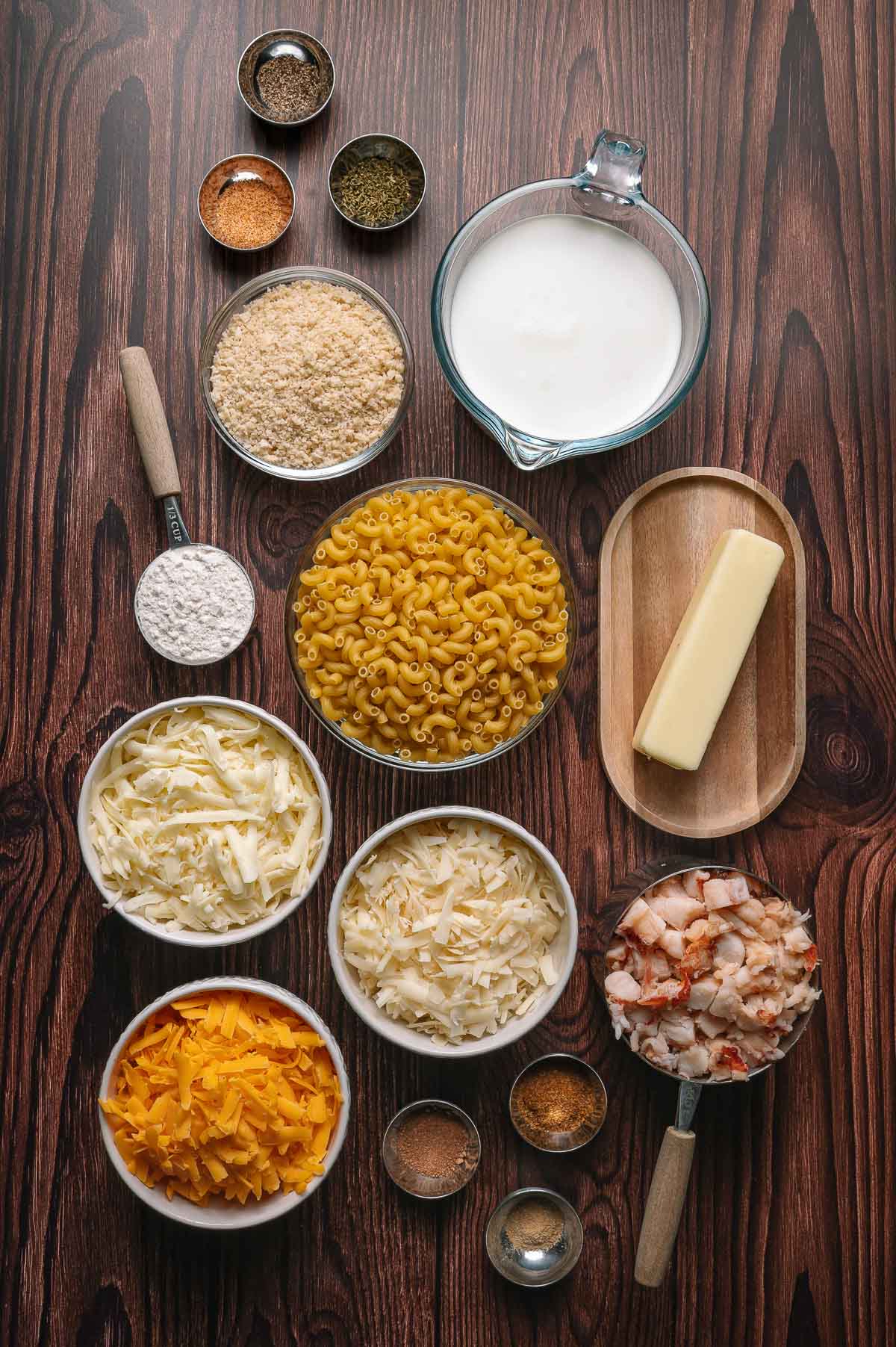 Ingredients for macaroni and cheese with lobster arranged on a wooden table, including pasta, shredded cheeses, milk, butter, flour, spices, breadcrumbs, and cooked lobster meat.
