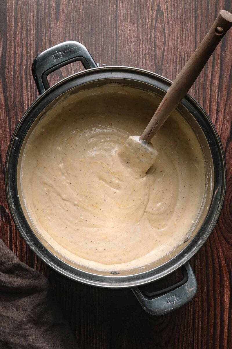 A metal mixing bowl filled with thick, smooth cheese sauce and a spatula resting inside, placed on a dark wooden surface.