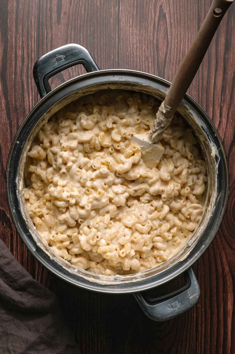 A pot filled with creamy macaroni and cheese sits on a wooden surface, with a spatula resting inside.