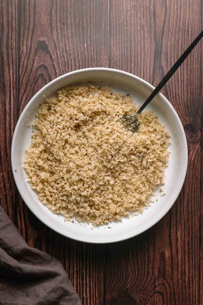 A white bowl filled with the macaroni and cheese panko topping on a wooden surface, with a spoon resting inside the bowl.