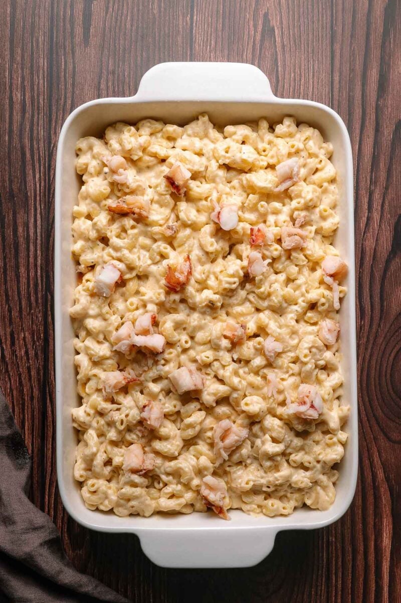 Rectangular white baking dish filled with macaroni and cheese, topped with pieces of lobster, on a wooden surface.