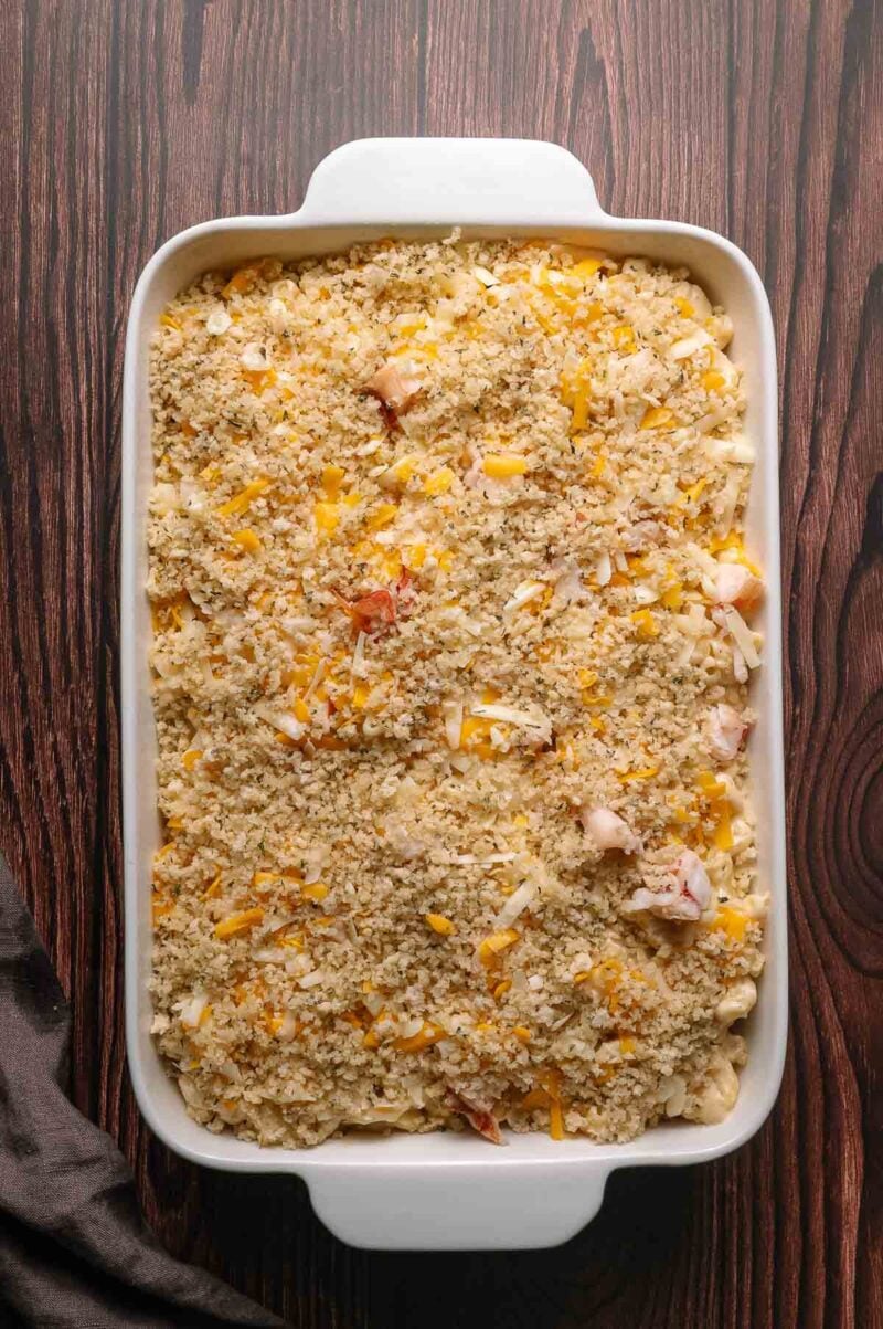 Rectangular white baking dish filled with macaroni and cheese topped with breadcrumbs, lobster meat and shredded cheese, resting on a wooden surface.