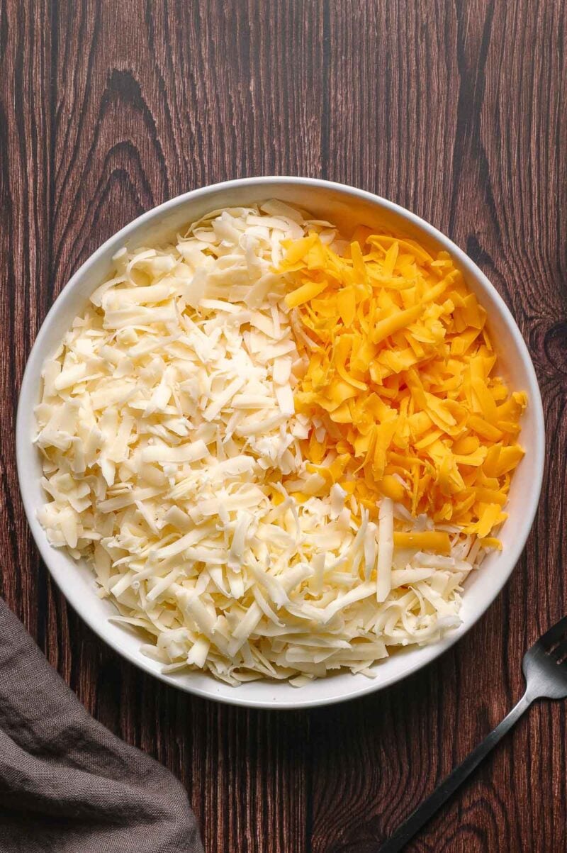 A bowl containing shredded white and yellow cheese sits on a wooden surface, with a fork and a cloth napkin nearby.