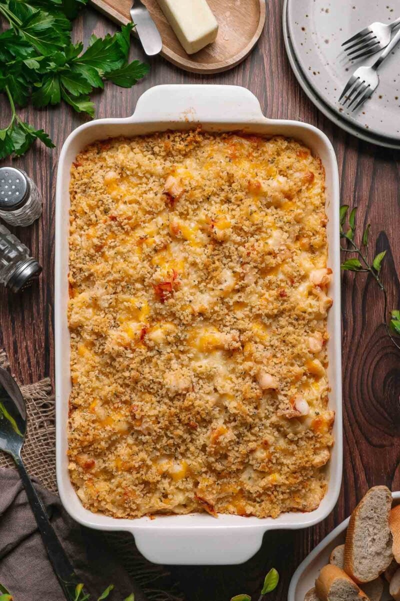 A baked lobster macaroni and cheese casserole with a golden breadcrumb topping in a white rectangular dish, shown on a wooden table with plates, cutlery, and bread nearby.