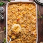 A baking dish filled with lobster mac and cheese topped with breadcrumbs; a serving spoon lifts a portion. Plates, utensils, and bread are nearby on a wooden table.