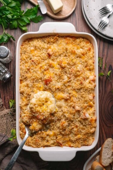 A baking dish filled with lobster mac and cheese topped with breadcrumbs; a serving spoon lifts a portion. Plates, utensils, and bread are nearby on a wooden table.